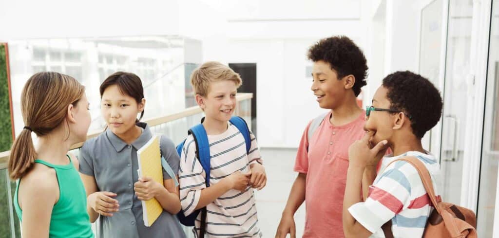 Diverse group of students talking and laughing in a school hallway, holding books and bags.
